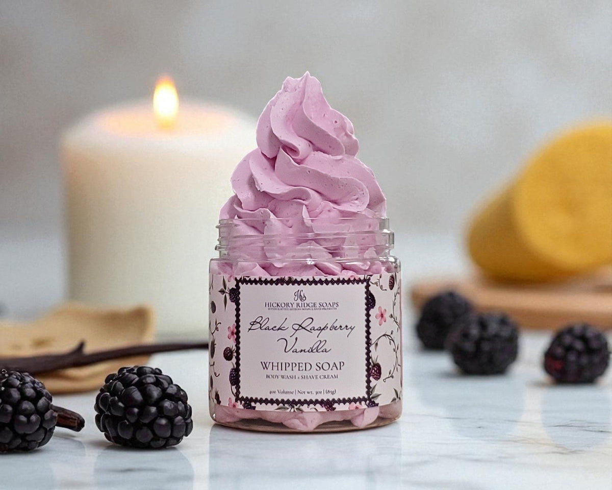 Pink whipped soap in a jar with blackberries and vanilla beans on a marble surface.