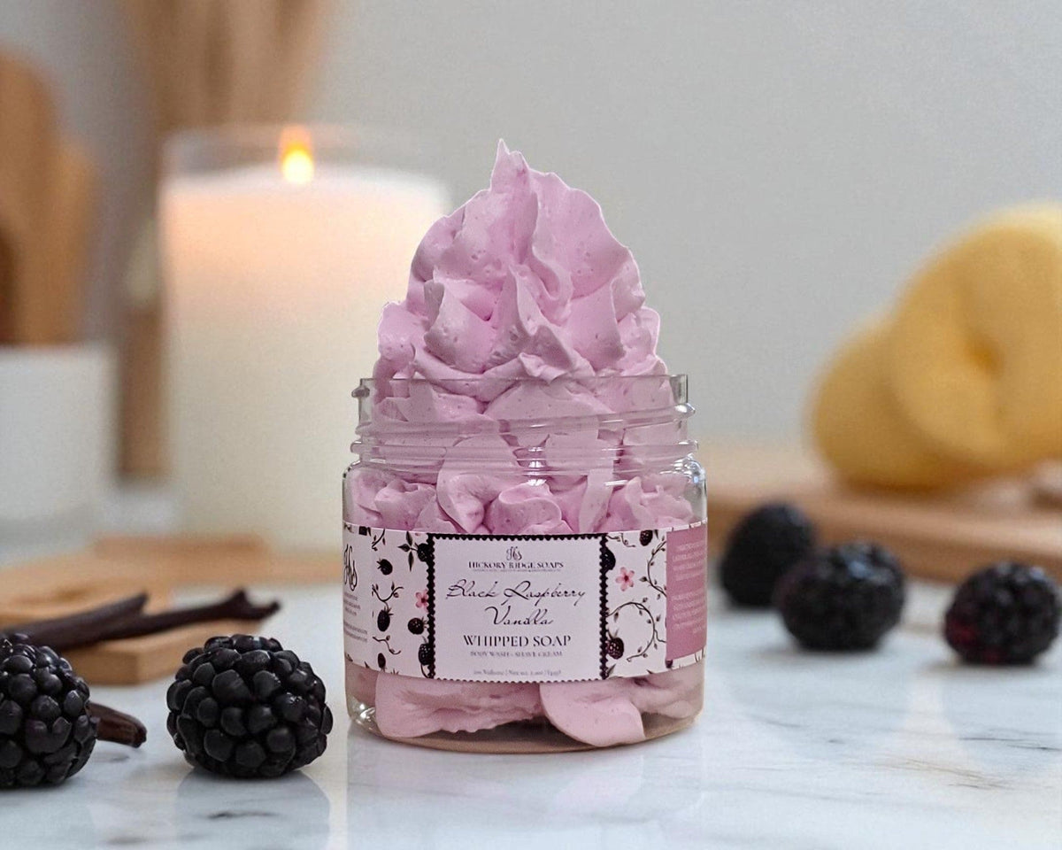 Jar of pink whipped soap with blackberries and vanilla beans on a marble surface.