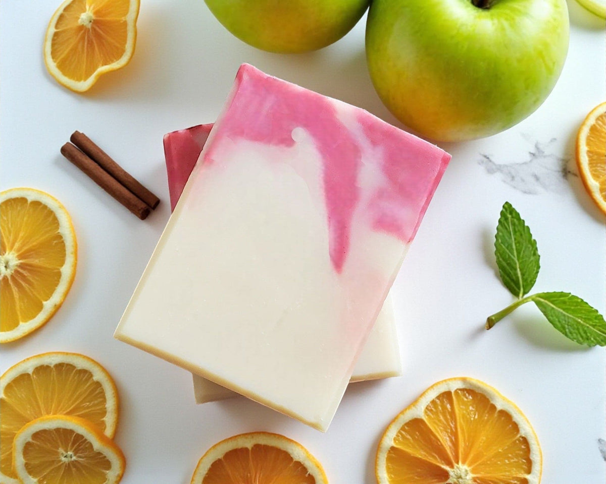 Two bars of soap with pink and white gradient surrounded by sliced oranges, apples, and cinnamon sticks on a marble surface.
