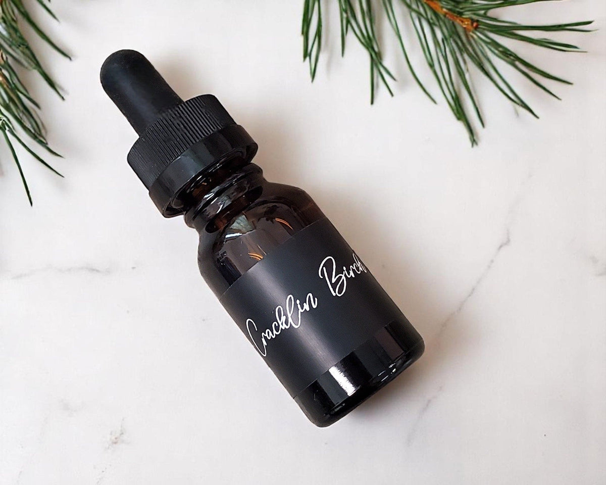 A bottle of cracklin birch car diffuser fragrance refill lying on a white marble surface.  There are pine branches in the upper part of the image.