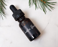 A bottle of cracklin birch car diffuser fragrance refill lying on a white marble surface.  There are pine branches in the upper part of the image.