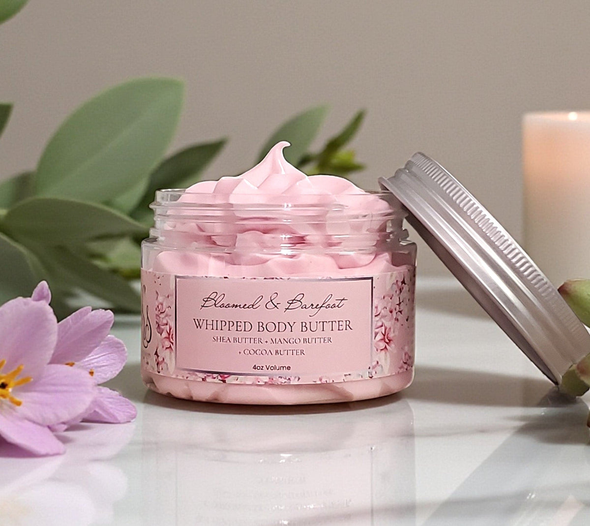 Jar of pink whipped body butter with a lid off, surrounded by flowers and leaves on a light surface.