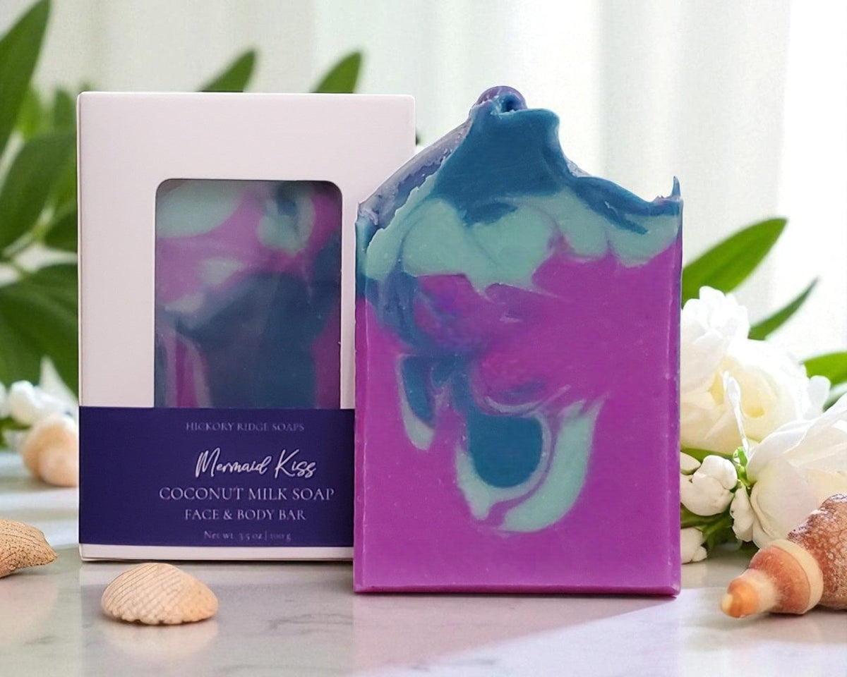 Purple and blue marbled soap bar with packaging on a white surface with greenery and shells.