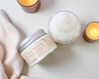 Two jars of whipped body butter with a towel and candles on a light surface