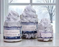 Three jars of 'Winter Wonderland' whipped soap on a windowsill with a snowy outdoor view.