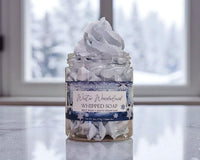 Jar of whipped soap with a label on a reflective surface, blurred window in the background